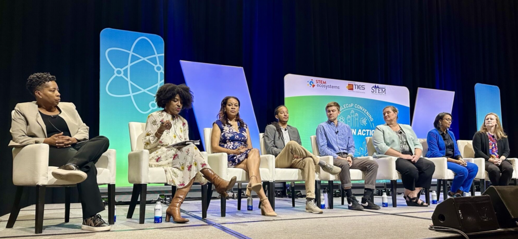 Panel discussion at STEM Ecosystems conference featuring eight diverse speakers seated on stage with blue and green backdrop displaying conference branding and atom symbol graphics.